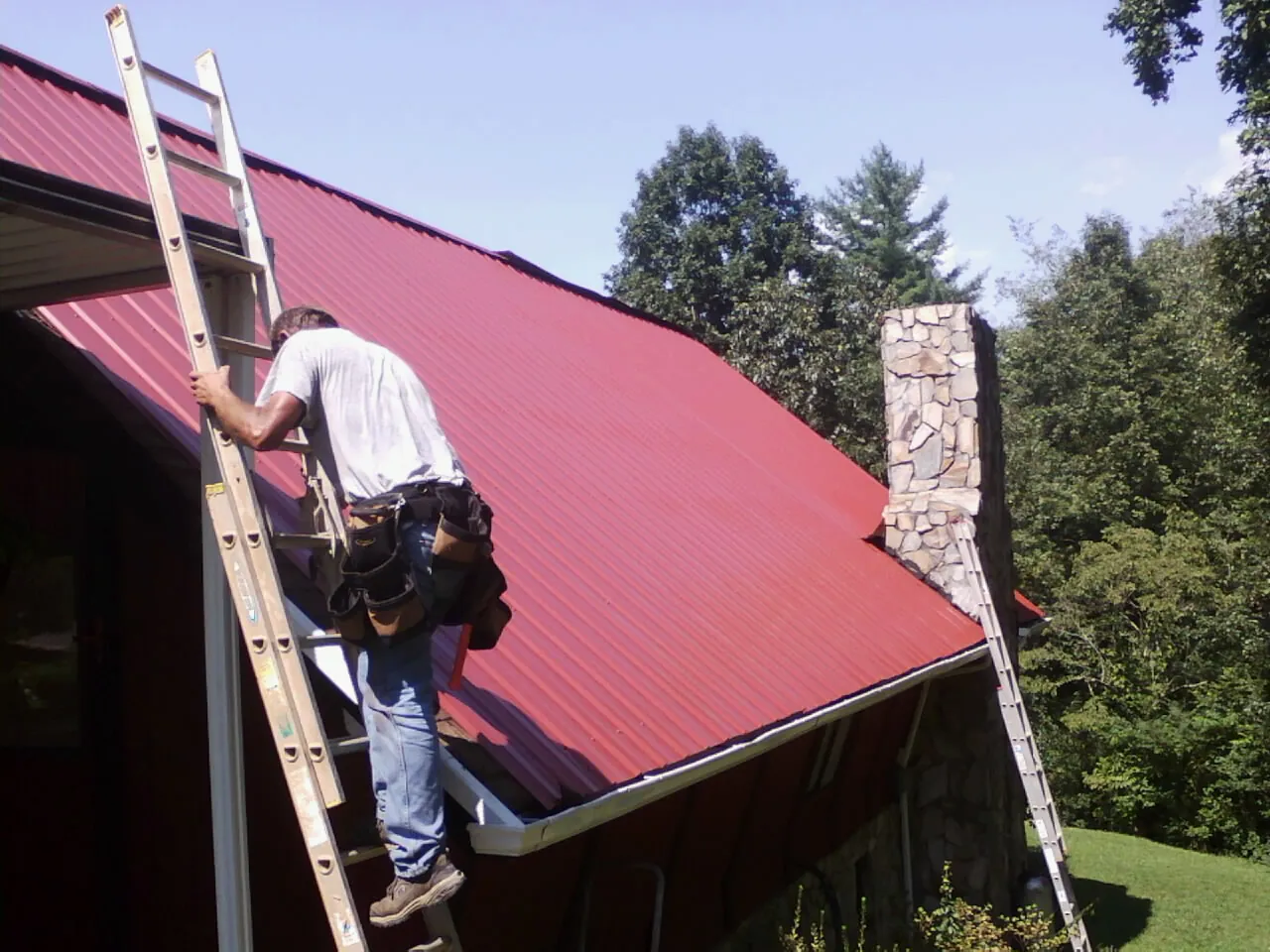 Roofer on a ladder beside a red metal roof and stone chimney — Soffit & Fascia Repair by Roofing Brothers in Mc Henry